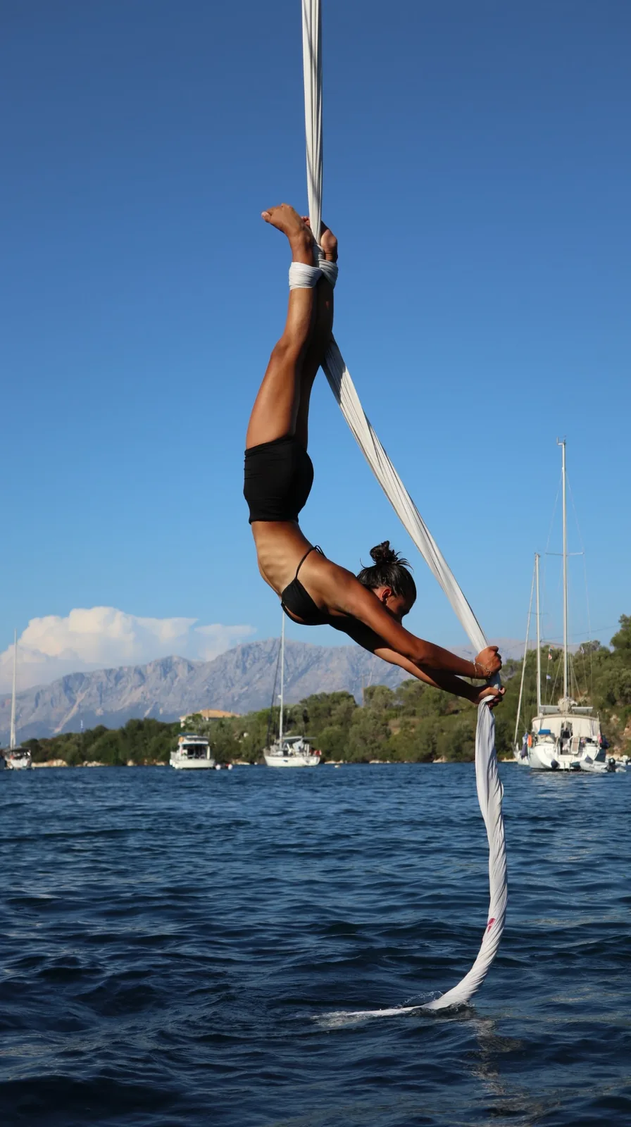Anaïs Badina inverted on white silks above a bay, moored boats and mountains behind